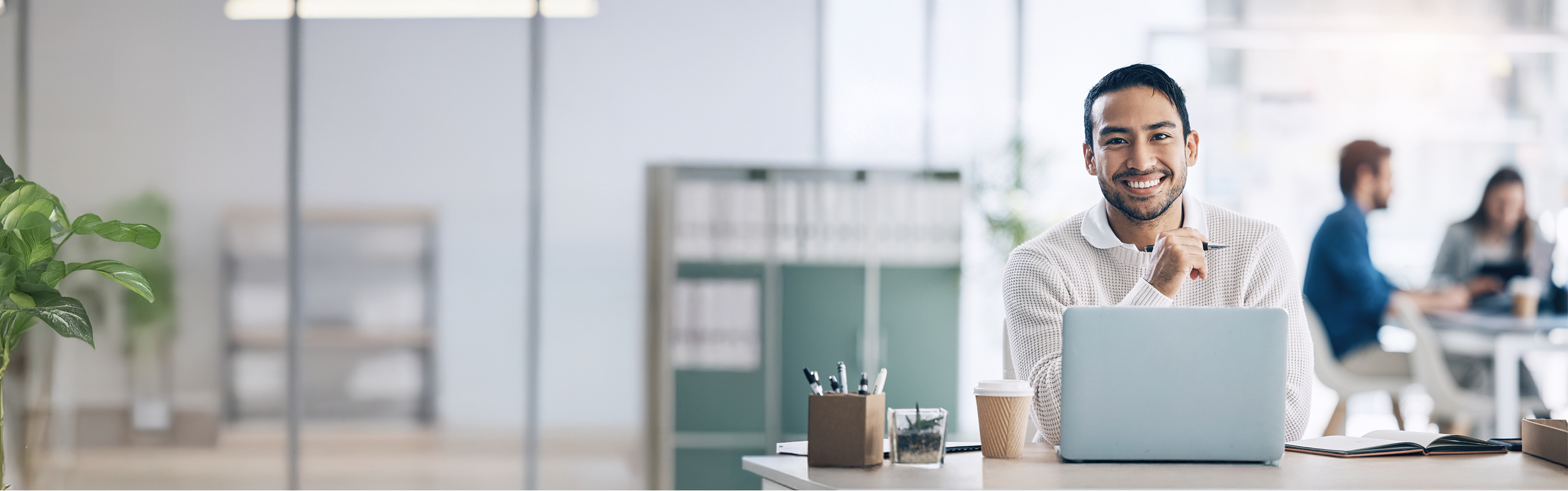 Komm ins Team von elio – Deine Karriere im Design und Marketing startet hier! Ein Mann sitzt an einem Schreibtisch in einem modernen Büro und arbeitet an einem Laptop. Im Vordergrund befinden sich Büromaterialien und ein Kaffeebecher. Im Hintergrund sind weitere Personen zu sehen, die ebenfalls arbeiten.