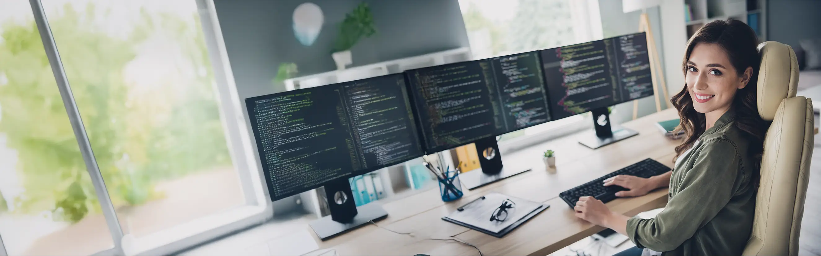 Komm ins Team von elio – Deine Karriere in der Entwicklung startet hier! Moderne Arbeitsumgebung mit drei Monitoren, auf denen Code angezeigt wird, und einer lächelnden Entwicklerin am Schreibtisch.