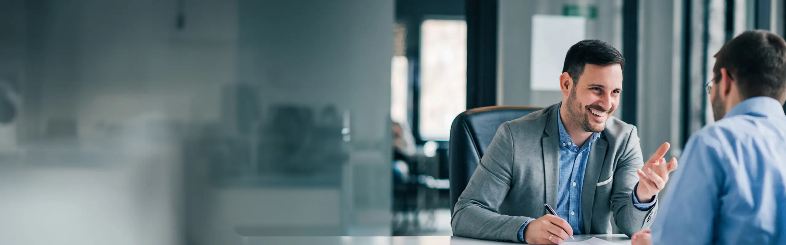 Digitalisierungsberatung Zwei Männer in einem modernen Büro im Gespräch – einer lächelnd mit Stift in der Hand, der andere mit dem Rücken zur Kamera.